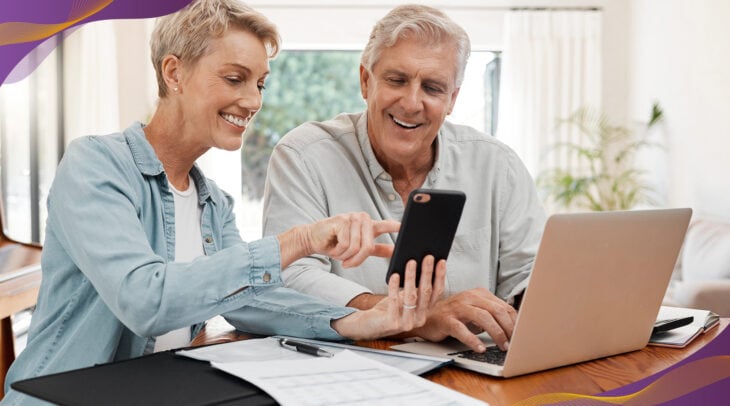 Older couple sitting at kitchen table looking at phone and computer.