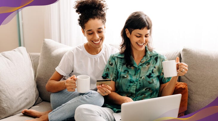 Woman and woman couple sitting on couch looking at laptop and drinking coffee