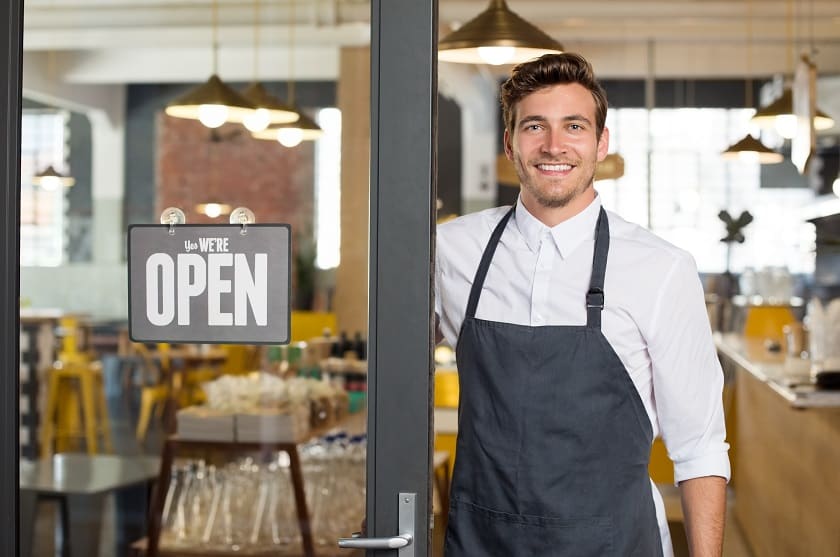 Smiling business male owner stands next to We're Open sign at front of restaurant