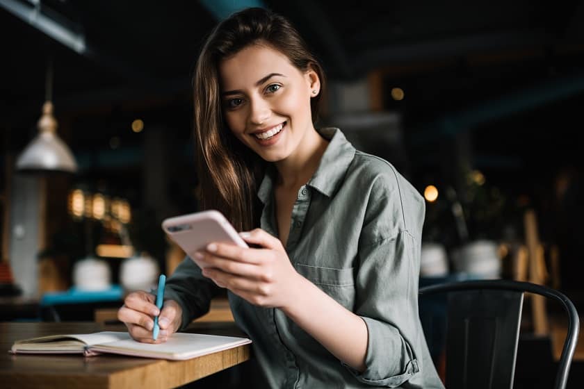 Smiling young woman uses cell phone and notepad and pen