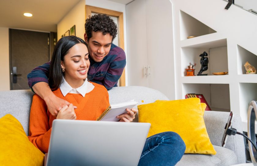 A young couple sits on a couch, looking at a notebook and a computer.