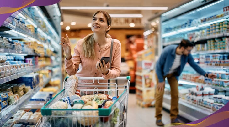 Adult woman shopping at grocery store pushing cart of items and holding cell phone.