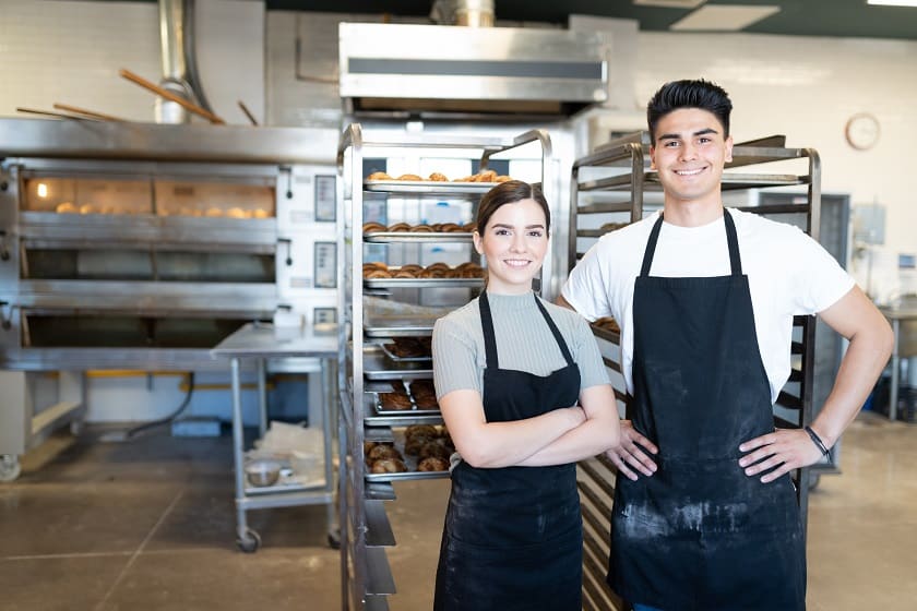 Young man and young woman wearing aprons stand next to bakery equipment
