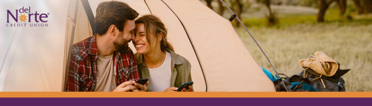 Man and woman on a date camping sitting outside tent looking at smartphones together.