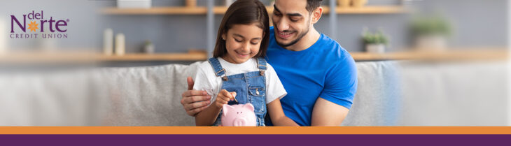 Father and daughter happy sitting on living room couch putting money into piggy bank.