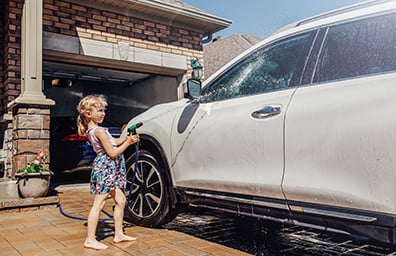 A little girl sprays a water hose at a white SUV in her family's driveway