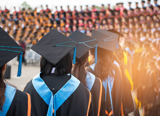 A line of graduating students with large group of onlookers walks during ceremony