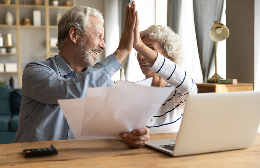 Older couple high five each other in front of laptop while holding bills