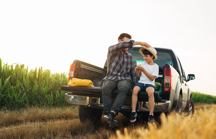 A man and his son sit in the bed of a truck in a field, and dad puts a hat on his son's head.