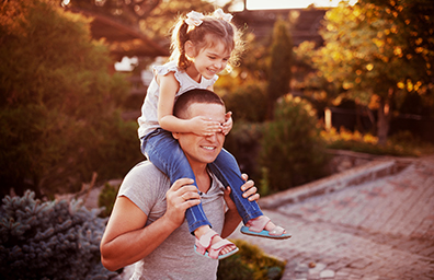 Smiling daughter sits on father's shoulders and covers his eyes with her hands