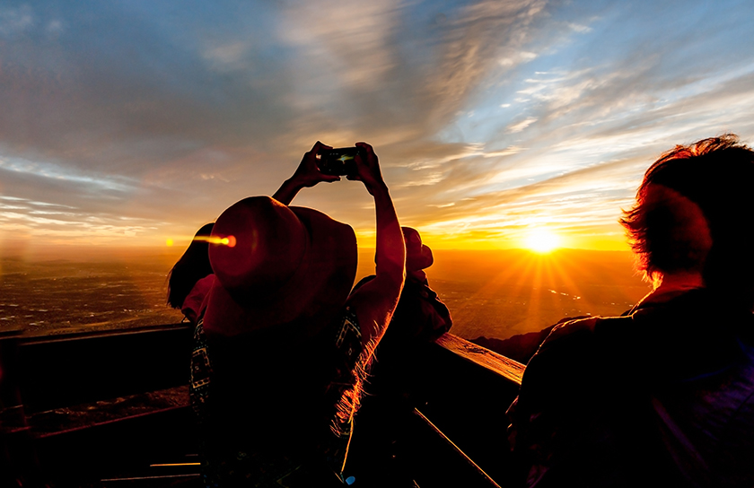 A woman with camera stands with two other people at scenic overlook during sunset