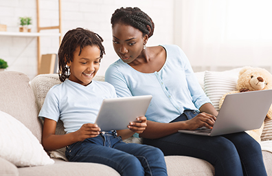 African American mother and daughter use electronic devices while sitting on couch