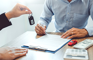 Man signs vehicle purchase documents at table while dealer hands car keys to him