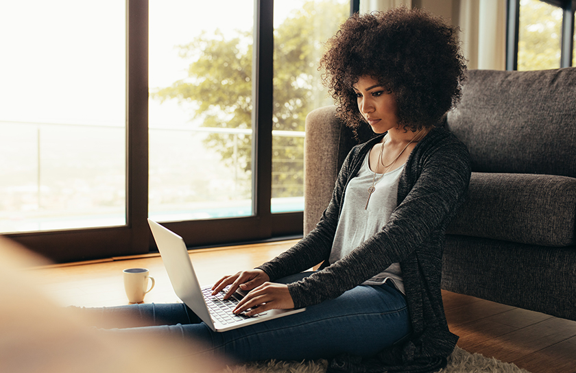 Woman sits on floor with back against couch and uses laptop