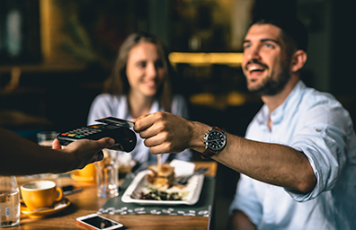 Smiling young man on date with woman hands credit card to waiter for payment